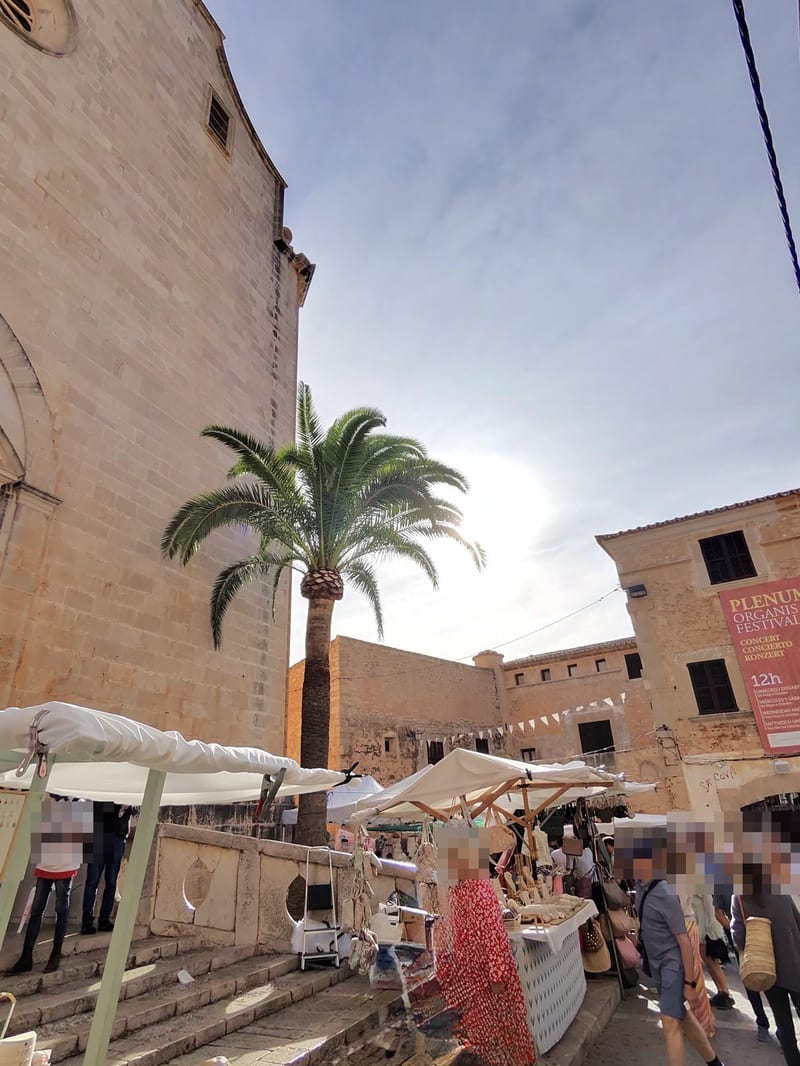 Kirche Sant Andreu in Santanyí mit hoher Palme, davor Marktstände mit Körben und Taschen, rechts ein Plakat des Plenum Organis Festival