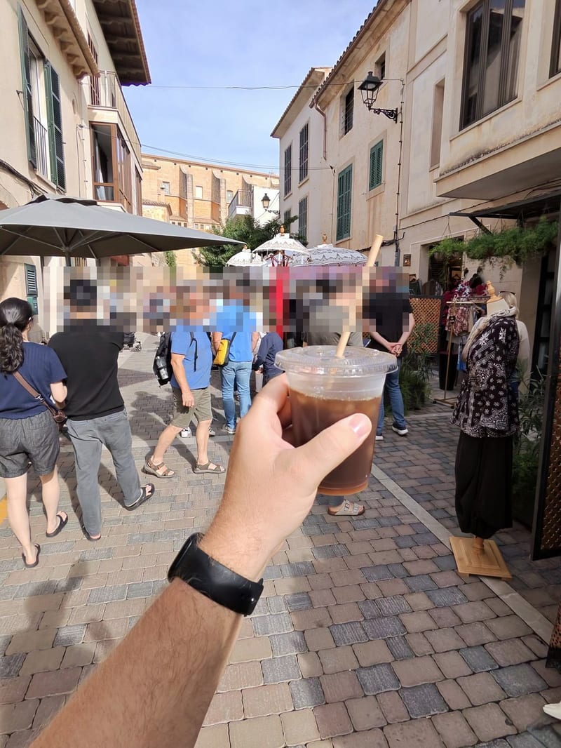 Hand mit Eiskaffee im Plastikbecher vor einer belebten Marktgasse