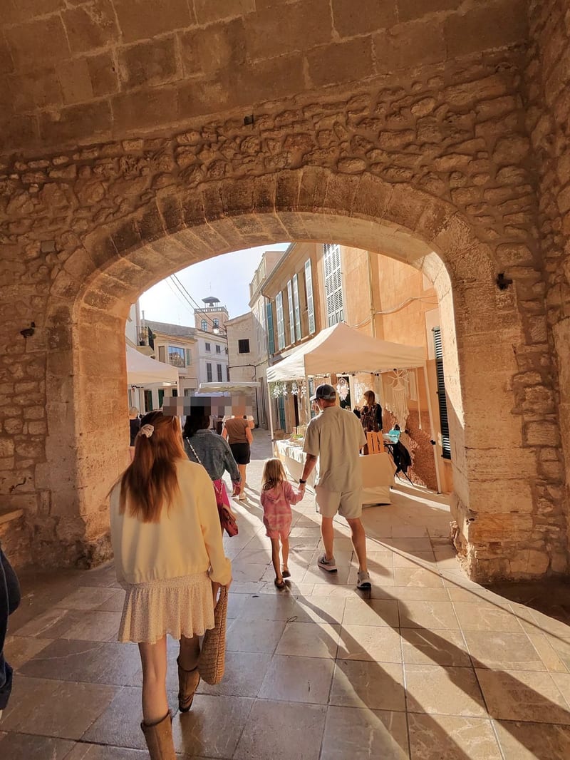 Durchgang unter einem alten Steinbogen, dahinter Gasse mit weißen Marktständen und Besuchern von hinten