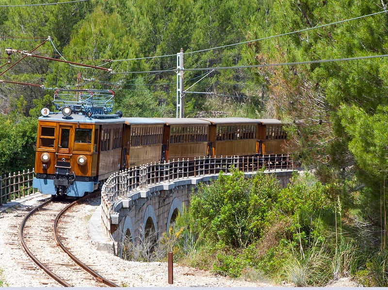Der Holzzug des Tren de Sóller fährt über eine Steinbrücke am Hang zwischen Pinien kurz vor Sóller