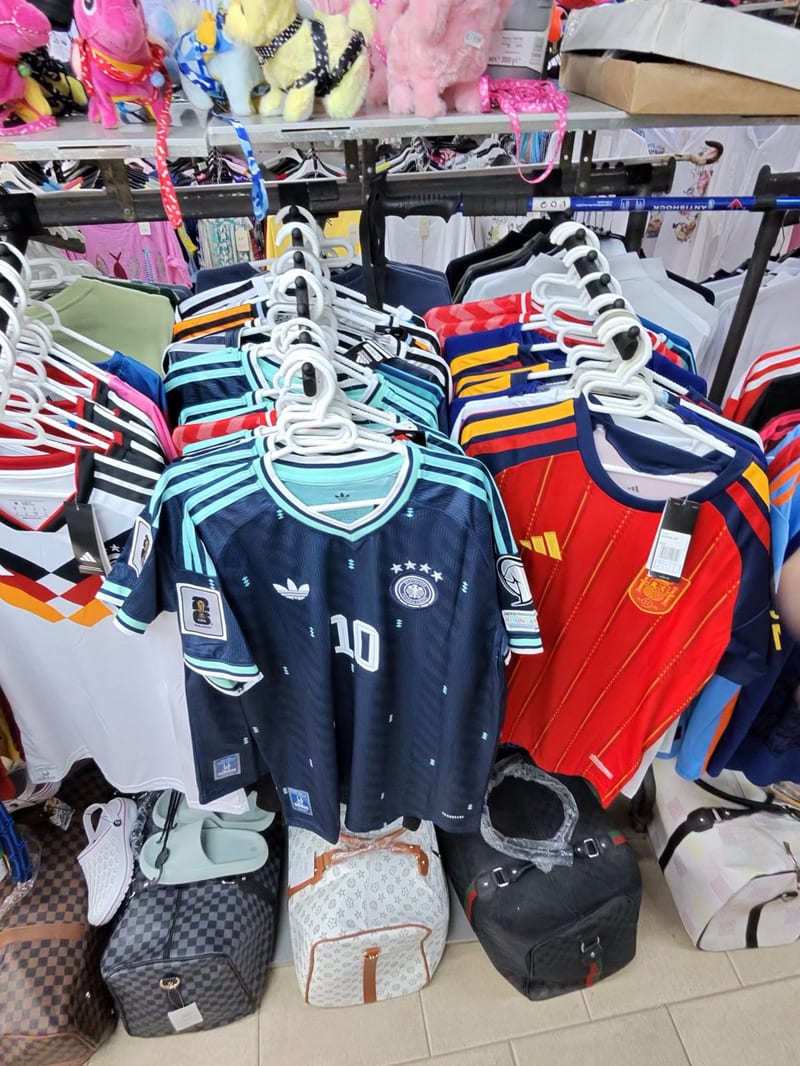 Kleiderständer in einem Laden auf Mallorca mit Fußballtrikots, vorn ein dunkelblaues Deutschland-Trikot mit der Nummer 10 und ein rotes Spanien-Trikot, darunter Reisetaschen