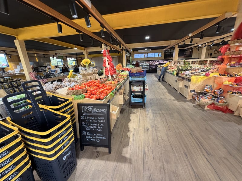 Innenraum von Agromart Balear in Inca: Holzstände mit Tomaten, Obst und Gemüse, vorn Einkaufskörbe und eine Kreidetafel der Theke „Ben trempat“