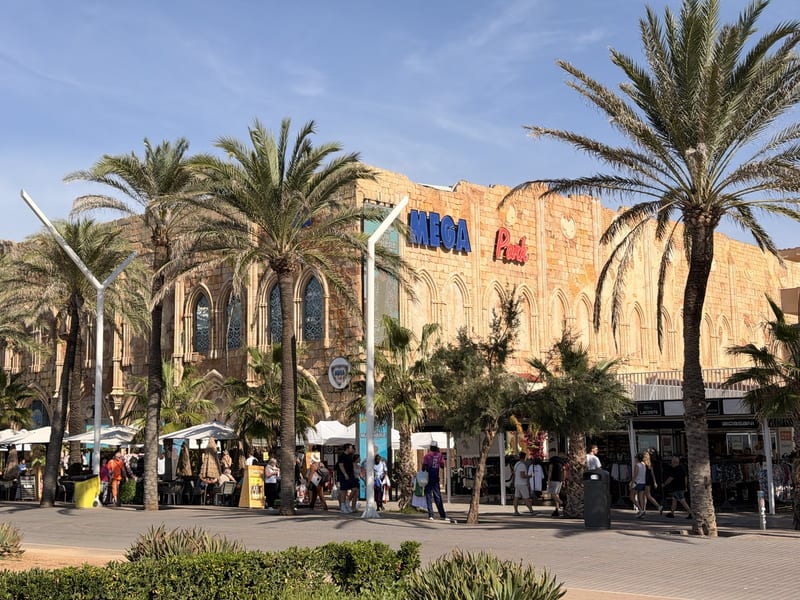 Fassade des Megapark an der Playa de Palma mit blau-rotem „Mega Park“-Schriftzug, Palmen und Terrassen bei Sonnenschein, davor Passanten auf der Promenade
