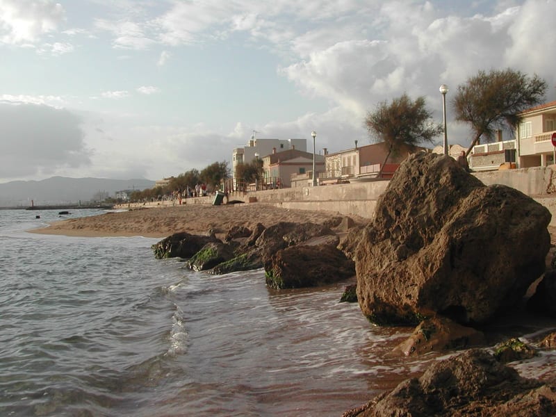 Felsen und kleiner Sandstrand an der Promenade von Es Molinar mit niedrigen Häusern