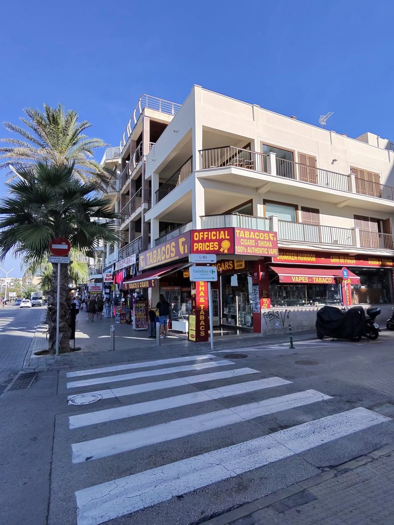 Eckgeschäft in S'Arenal mit gelb-roten Schildern „Tabacos“, „Official Prices“, „Cigars – Shisha“ und „Vapes – Tabaco“ unter einem weißen Apartmenthaus, davor ein Zebrastreifen, Palmen und blauer Himmel
