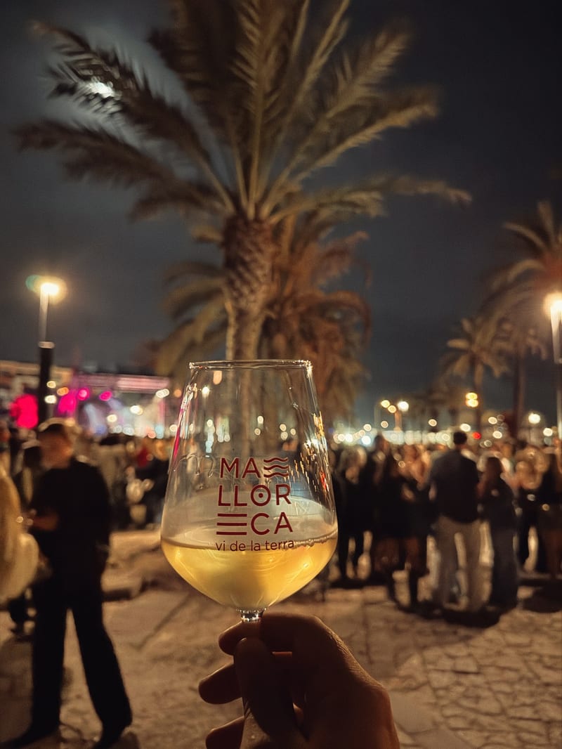 Weinglas mit Weißwein und dem Aufdruck Mallorca vi de la terra vor Palmen und Festbesuchern bei Nacht